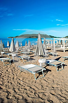 Sun loungers and umbrellas on public beach at summer sunny mornin. Skojl island background. Calm sea and blue sky Becici, photo