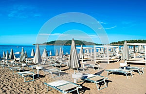 Sun loungers and umbrellas on public beach at summer sunny mornin. Skojl island background. Calm sea and blue sky Becici, photo