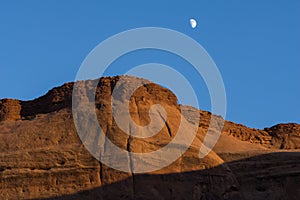 Sun lowers in sky and brightens a red rock escarpment under white moon