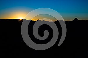 Sun rising over black foreground with jagged desert plateau and mesas silhouetted against the blue sky