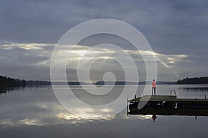 The sun`s rays make their way through the clouds above the lake, a female silhouette on a wooden pier