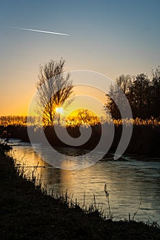 The sun sets behind a tree over the dutch countryside. The water in the canal is frozen and reflects the colors of the sky.