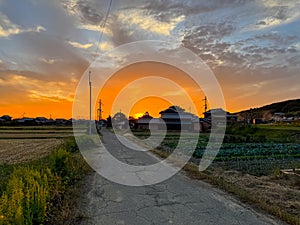 Sun sets by country road and rural Japanese houses under dramatic sky