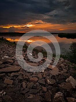 The sun sets on a river bank filled with rocks and also surrounded by dense forests and vast land