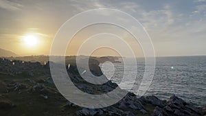 The sun is setting over the ocean, casting a warm glow over the rocky shoreline