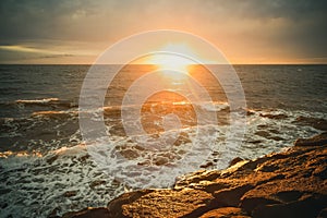 Sun setting over water with waves and rocks.