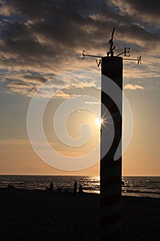 The sun is setting on the sea from behind a pillar with a wind rose