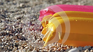 Sun shines to yellow and pink coloured inflatable with drops on water at surface, small pebbles beach under, closeup detail,