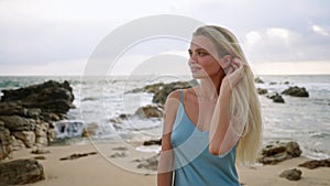 Sun-tanned woman in blue dress enjoys serene beach. Blonde hair, blue sea, stroll on sandy shore. Leisure, travel