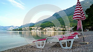Sun umbrellas and loungers on empty beach with town and mountains on background
