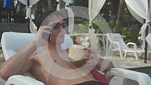 Sunbathing tanned man wearing sunglasses with bare chest talking on cellphone by the sea and drink coconut juice