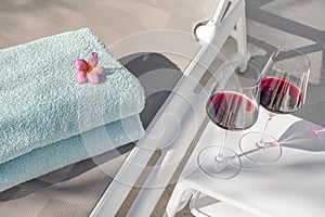 Sunbed with two towels neatly folded and a white table near it with two tall glasses of red wine, poolside inviting setting