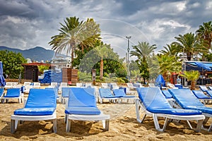 Sunbeds on sand on tropical beach resort in bad weather with cloudy sky. Palm trees and chaise lounges on beach in cloudy weather.