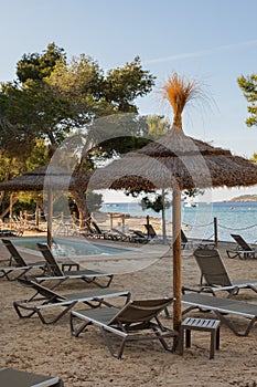 Sunbeds and thatched sunshades on the beach of a hotel with swimming pool and view to the sea on Ibiza island. Beautiful