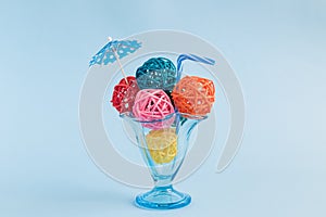 Rattan balls in form of ice cream scoops with paper parasol and drinking straw in glass cup on blue background minimalistic concep