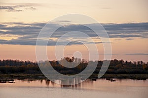 Sundown and sunrises. Blue sky with tints of purple and much clouds. Silhouettes of trees and grass are reflectioning in the river