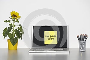 Sunflower plant on desk and sticky notepaper on laptop screen with fare qualche lavoro written on it in Italian