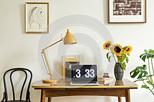 Sunflowers, yellow lamp and laptop on wooden desk in home office interior with posters. Real photo