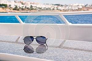 Sunglasses lie on a table on a white yacht against the background of the sea and the shore.