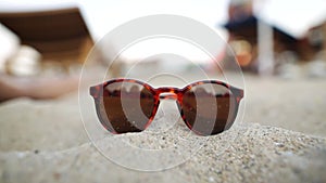 Sunglasses lying on the sand. Unrecognizable people, sandy beach, chasers and the sea are on a blurred background.