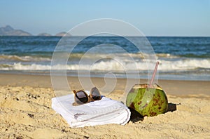 Sunglasses with towel and a coconut on the sandy beach with blurry Atlantic ocean in backdrop