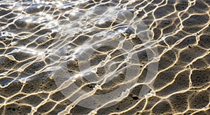 Sunlight creates intricate patterns on a sandy surface beneath shallow water. The light refracts