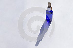 Sunlit blue galss bottle with pipette full of skin care serum on white background with long shadows.