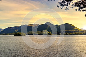 Sunlit Derwentwater lake surface with a greeny mountain range behind seen through tree branches