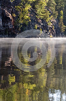 Sunlit Forested Cliffs reflected in still misty water