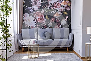 Sunlit, gray sofa by a floral print wall in the nook of a feminine living room interior with golden accessories