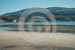 Sunlougers below umbrellas in the shallow water of turquoise bay. Opposite mountainous island with windmills on the top