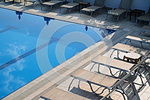 Sunlounger chairs near an outdoor pool and cloudy sky reflecting in the blue water