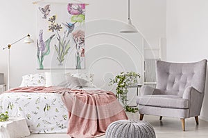 A sunny bedroom interior with a bed dressed in green pattern white linen and a peach blanket. Gray comfortable armchair beside the