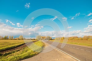 A sunny country road in early autumn or summer time with yellow and green grass, trees and blue sky with white clouds