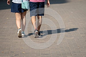 On a sunny day, a couple of people walk along the sidewalk. The human shadows are visible on the sidewalk. View from the back.