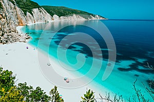 Sunny Fteri beach lagoon with rocky coastline, Kefalonia, Greece. Tourists under umbrella chill relax near clear blue