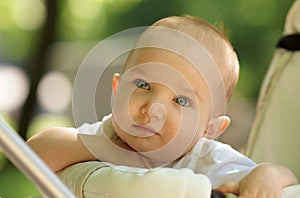 Sunny portrait of cute baby in white shirt sitting in baby carriage. Thoughtful baby with big blue eyes. Closeup
