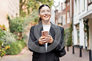 Sunny portrait of a newly entrepreneur young woman in her 25s, black suit, holding a phone smiling, looking away