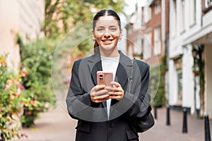 Sunny portrait of a newly entrepreneur young woman in her 25s, black suit, holding a phone smiling, looking in camera