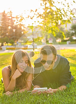 Sunny portrait of sweet young couple lying relaxing on the grass together listens to music in earphones on smartphone
