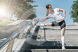 Sunny summer day. Young woman doing stretching exercises outdoor. Girl doing warm-up on steps before training.