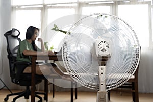 Sunny summer in front of the working fan, suffering from the summer heat. Unhappy Asian woman sitting in front of fan at work