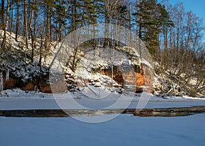 Winter landscape with red sandstone cliffs on the bank of the river Salaca, the sun shines on the trees and the river bank, the