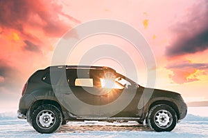 Sunrays Through Window Of SUV Car Parked On Winter Snowy Field At Sunset Dawn Sunrise. Altered Sunset Sky