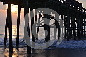 The sunrise appears through the beach pier pilings and reflects on the ocean waves