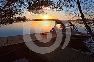 Sunrise on calm lake, floating pier with moored motorboat under pine branches, nobody