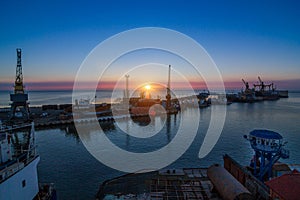 Sunrise illuminates sea port, cargo ships docked at berth. Industrial cranes against dawn sky. Freight containers ready