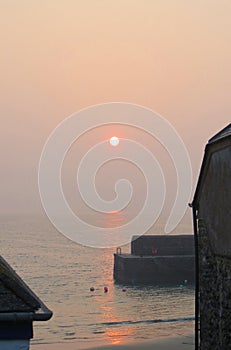 At sunrise a misty path of sunlight leads across the sea to the fishermen\'s net lofts at Gorran Haven quay Cornwall England
