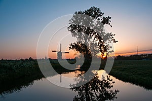 Sunrise with old tree and Dutch windmill reflected in the water
