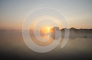 Sunrise over the foggy lake with the reflection of sun and trees in the water. Early fresh morning landscape. Mist on the water,